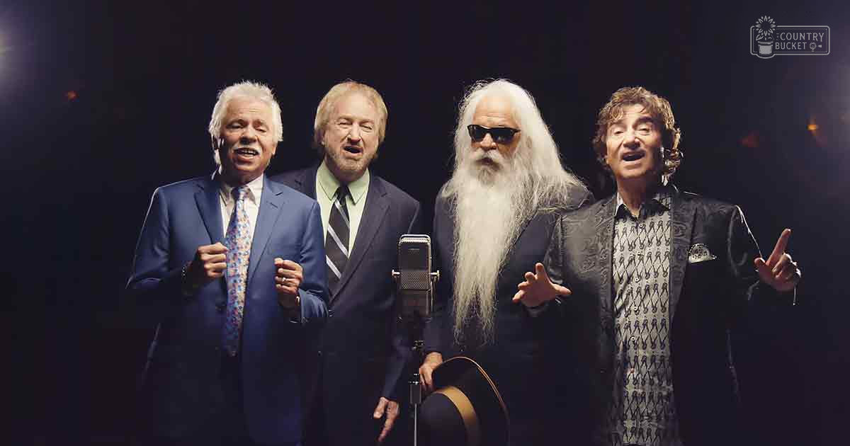 Performers on stage, older adult male in blue suit jacket with patterned tie, middle-aged male in dark suit with striped tie, older adult male with long white hair and sunglasses, middle-aged male with curly brown hair wearing a dark blazer. Microphone visible, stage lighting focused, indoor venue at evening.