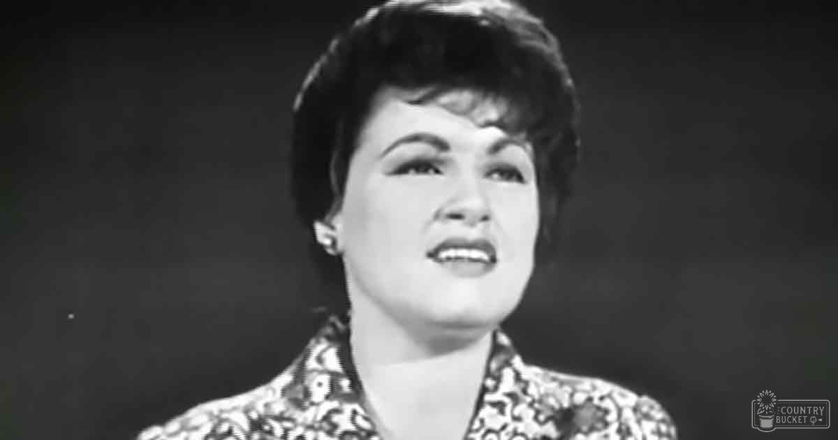 Middle-aged adult female wearing a patterned blouse with floral or paisley design in an indoor setting with dark background and spotlight effect, possibly on stage.