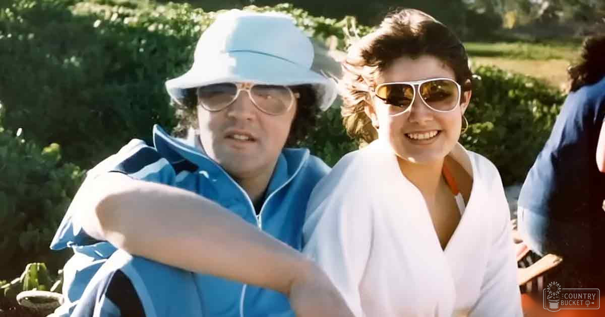 Adult males and a female outdoors on bright sunny day with greenery; man wears light blue jacket from 1980s style over dark shirt, white bucket hat, aviator sunglasses.