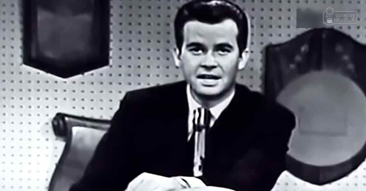 A young adult male wearing a dark suit, white dress shirt with vertical stripes, and a striped tie stands against an indoor mid-20th century office backdrop.