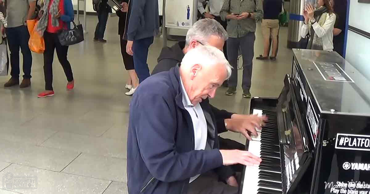 A man playing a piano on a train station platform with other people waiting in the background.