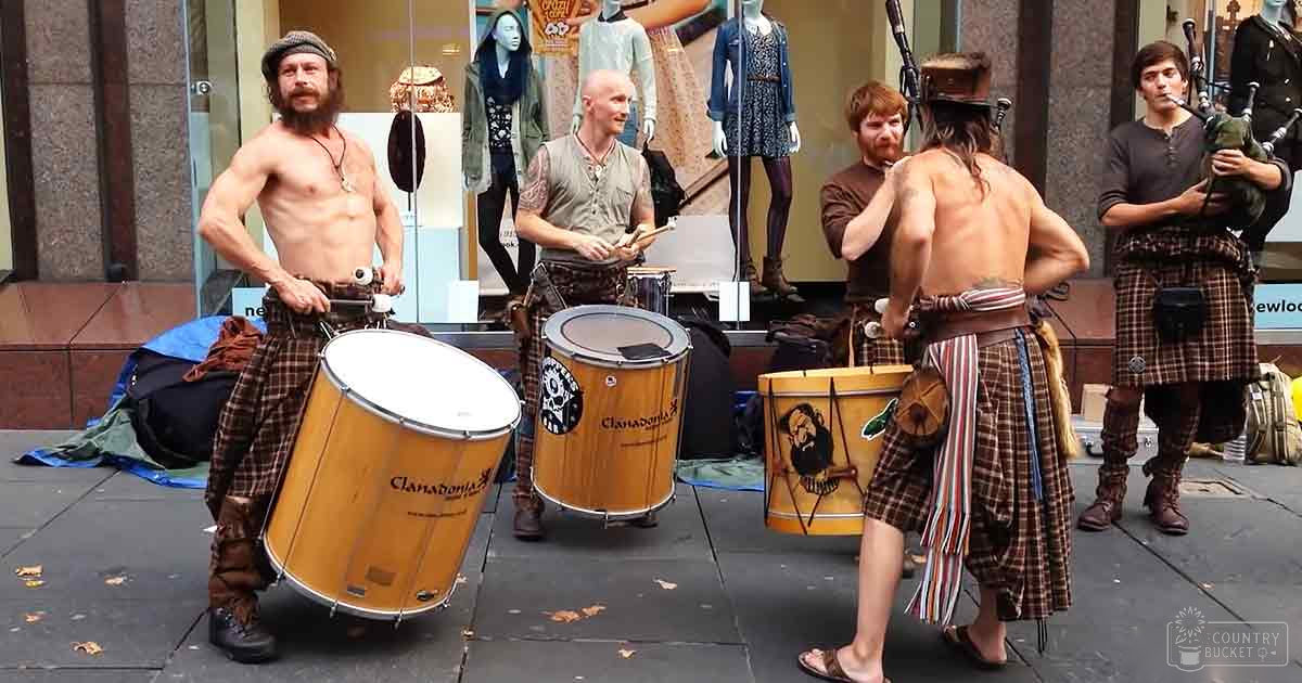 Street performance group in urban setting, adults wearing plaid skirt-like garments and various footwear; mannequins dressed in outfits displayed behind them.