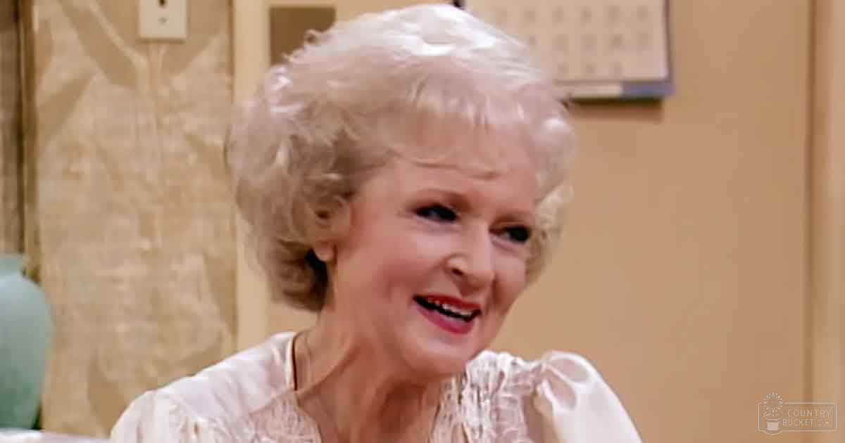 An older adult woman with short curly white hair wearing a light-colored ruffled blouse in an indoor domestic setting during daytime.
