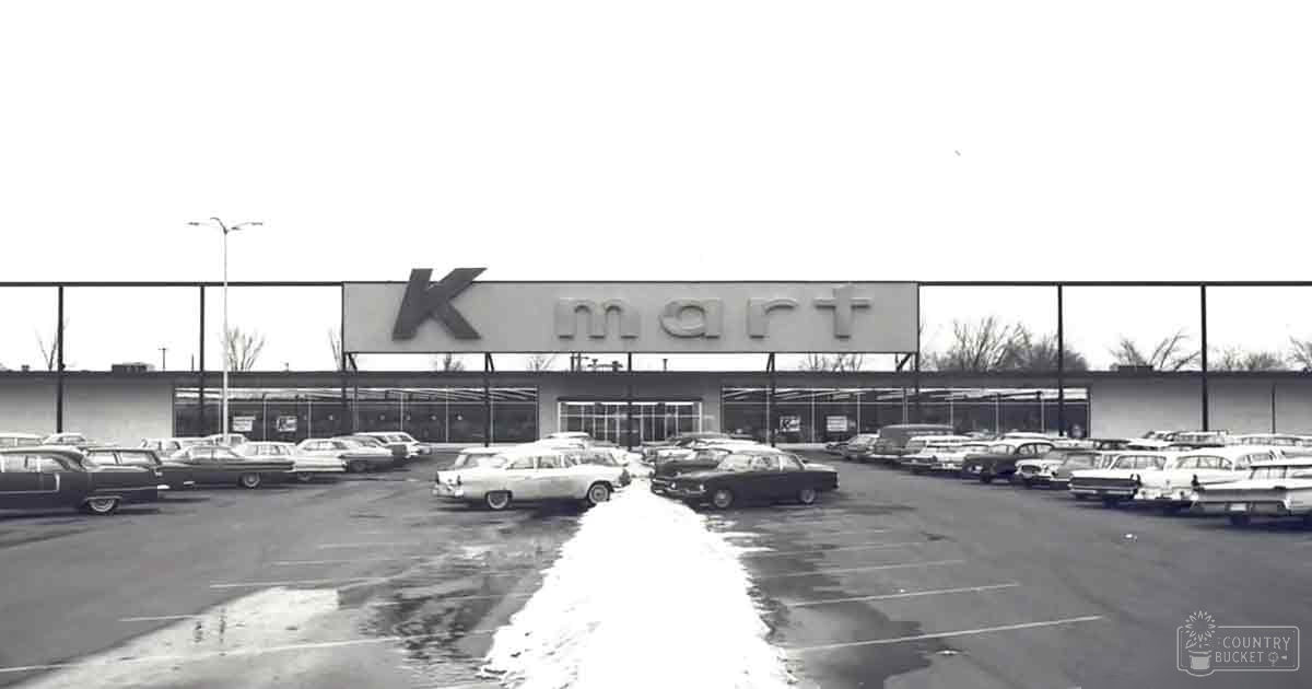 Vintage cars lined up in front of a Kmart store with bare trees and overcast skies suggesting late autumn or winter, black-and-white image reflecting mid-20th century design trends.