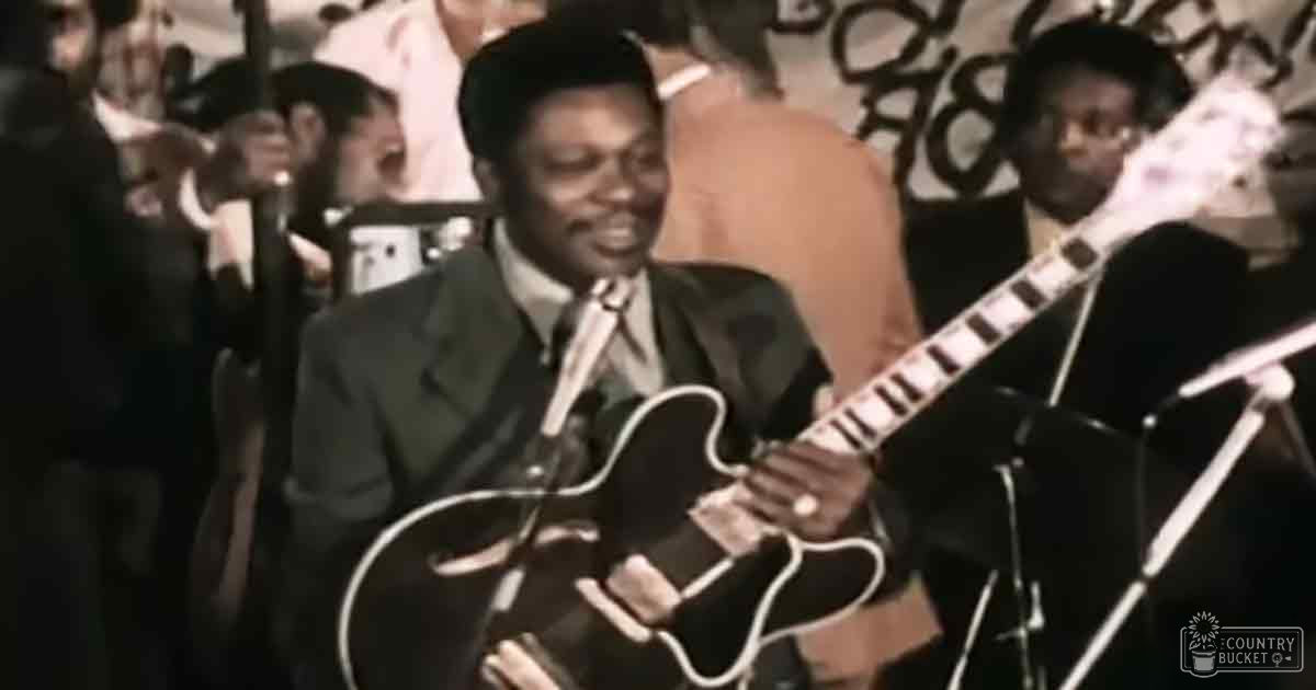 A mid-aged male musician in a dark suit jacket and white shirt playing guitar indoors with an audience behind, under stage lighting from what appears to be a concert setting typical of the mid-20th century.