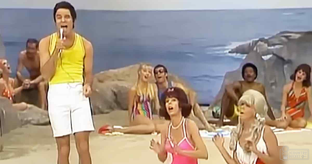 A group of women wearing swimsuits indoors, with an adult male singer holding a microphone against a backdrop with a mural of rocks and water.