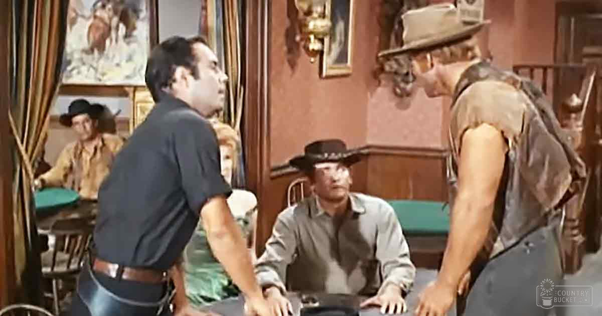 A scene from a Western movie or TV show set in an old saloon, with men dressed in period clothing seated around a bar. Wooden walls and rustic decor suggest late 19th-century to early 20th-century setting.