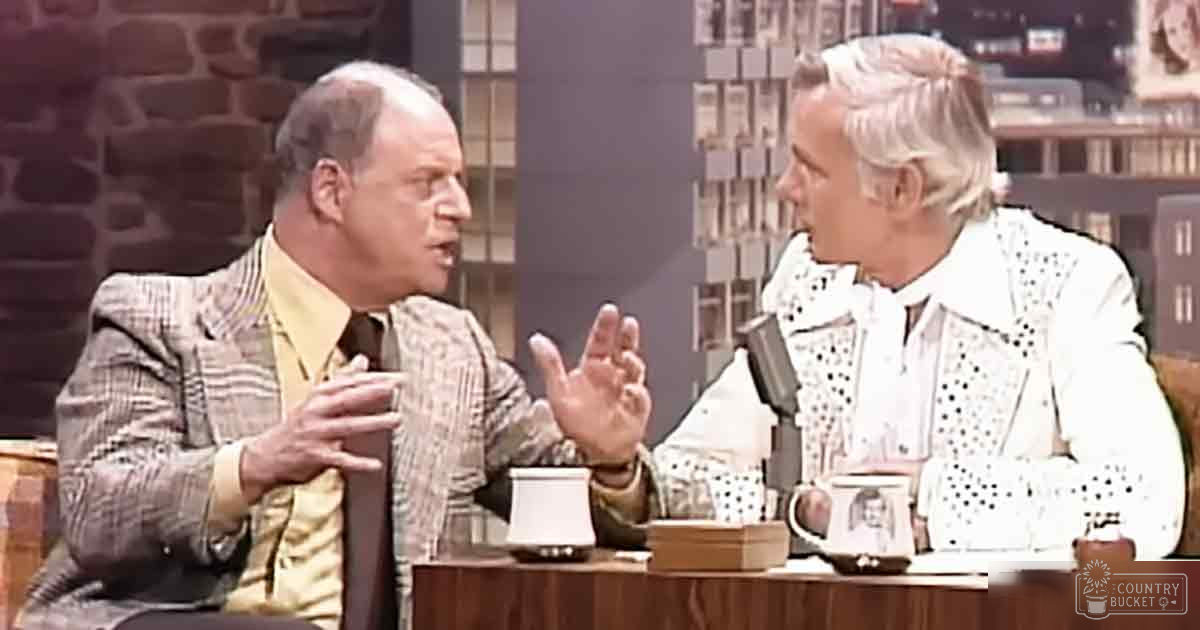 Two middle-aged to older adult men in a mid-20th century talk show setting, wearing formal and semi-formal attire with wood furniture and stone wall background.