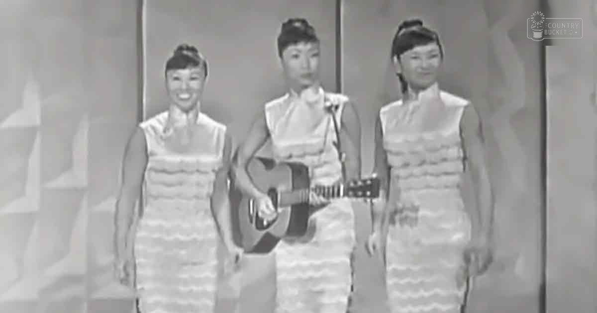 A musical performance scene in a studio setting with evenly lit, bright indoor lighting; three adult females wearing matching sleeveless dresses from the mid-20th century, positioned on stage.