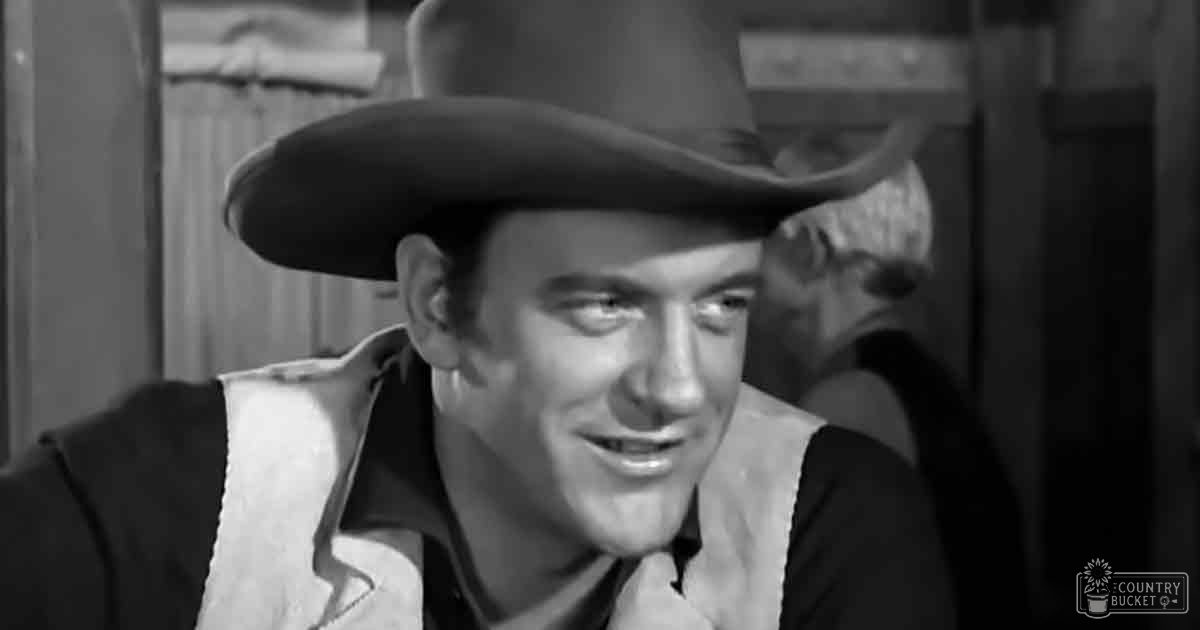 A young adult male wearing a wide-brimmed cowboy hat and Western attire stands indoors within what appears to be an old Western-style building, possibly a saloon or cabin.