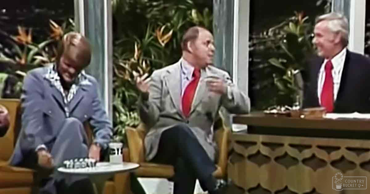 Talk show set with three adult male guests seated indoors, surrounded by decorative plants and large windows showing lush greenery outside, suggesting daytime in spring or summer; indoor studio lighting is bright but not harsh.
