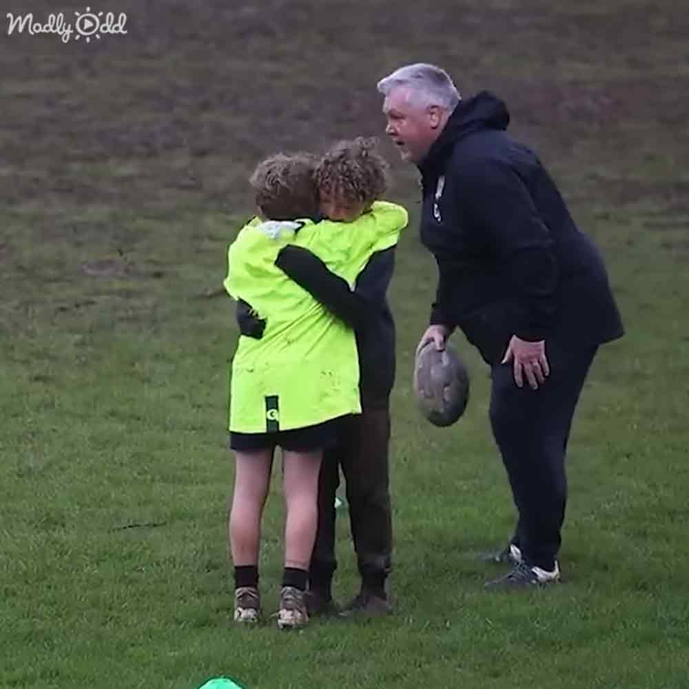 An outdoor sports field with an adult comforting a young child wearing a neon green jacket on a grassy field, likely autumn due to the bare trees in the background.