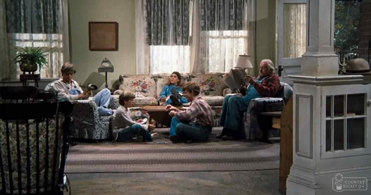 A family scene with children and adults sitting on a floral-patterned couch indoors, natural light streaming through large windows suggesting daytime in spring or summer.