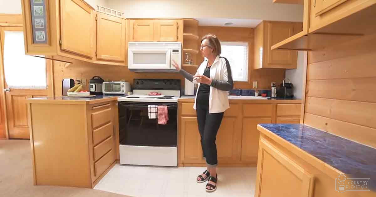 A middle-aged female stands in a well-lit kitchen, wearing a white cardigan over black top and pants, with sandals on her feet; wooden cabinets, white microwave, black stove, blue speckled countertop, red checkered towel hang from oven handle.