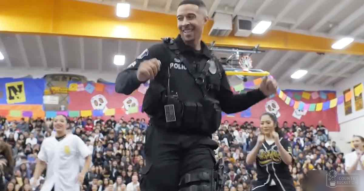 An adult male police officer in a black tactical uniform stands inside an indoor school gymnasium with bright artificial lighting and colorful banners with skulls. Children and adults are present, some in casual clothes.
