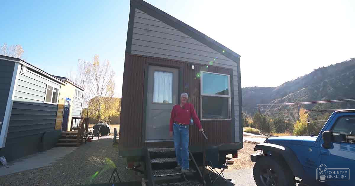 Middle-aged adult male wearing a red long-sleeve shirt, blue jeans, and dark shoes or boots stands in front of a tiny house with mountains in the background. The setting is a rural area with clear skies and autumn-colored trees.