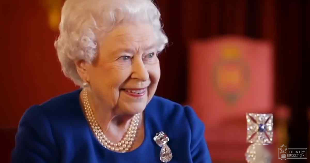 An older adult woman in a blue dress or jacket indoors, wearing a pearl necklace and a brooch with a teardrop-shaped gemstone, surrounded by red curtains.