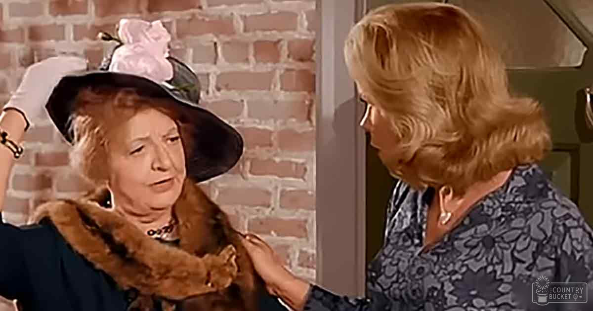An older adult woman wearing a black brimmed hat with pink floral decoration and a dark blue coat with a fur collar stands next to another middle-aged female in a patterned blouse indoors against a brick wall backdrop.