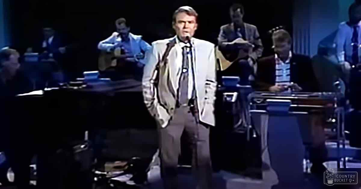 A middle-aged man in a white suit jacket sings on an indoor stage with an orchestra, illuminated by stage lights.