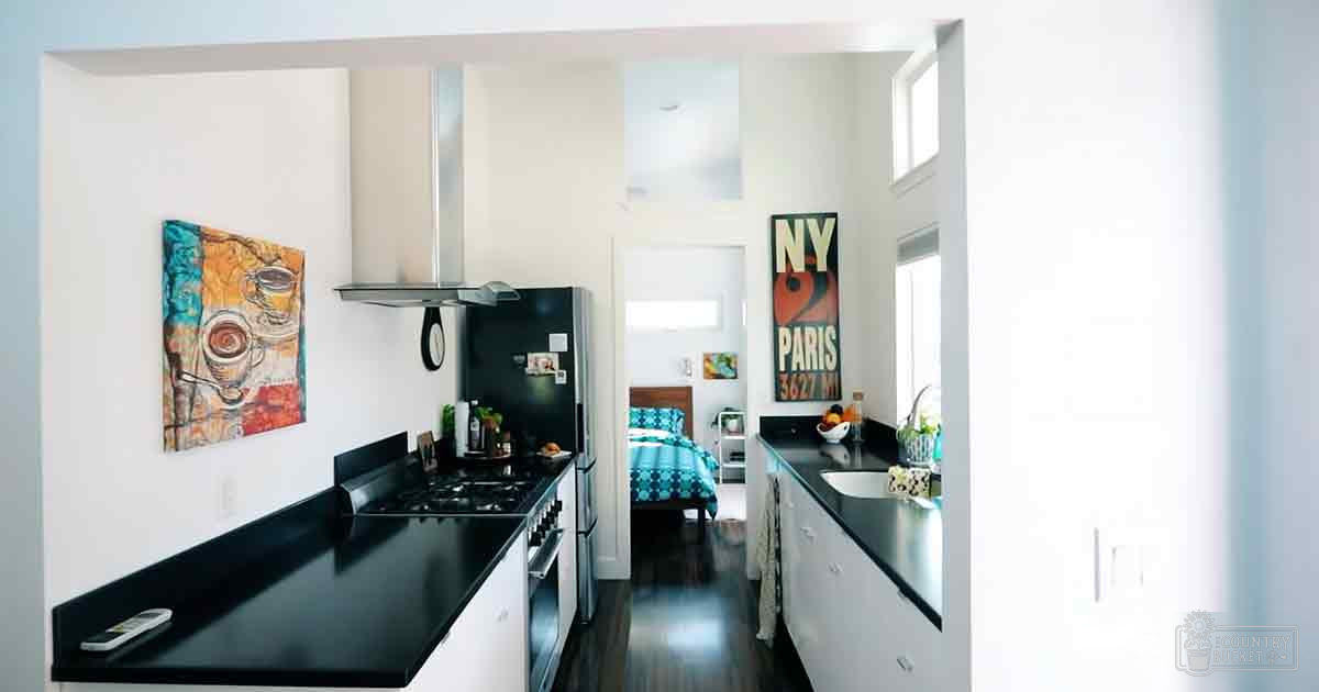A kitchen with a gas stove, stainless steel refrigerator, sink area, colorful coffee cup painting, and 'NY PARIS 3627 M' poster in an apartment with dark wooden flooring and natural light.