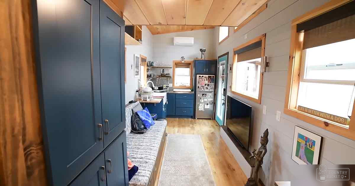 A cozy interior space with blue cabinets, a gray couch with patterned cushions and a blue blanket, wooden flooring, a small kitchen area, windows with wooden frames, and a flat-screen TV mounted on the wall.