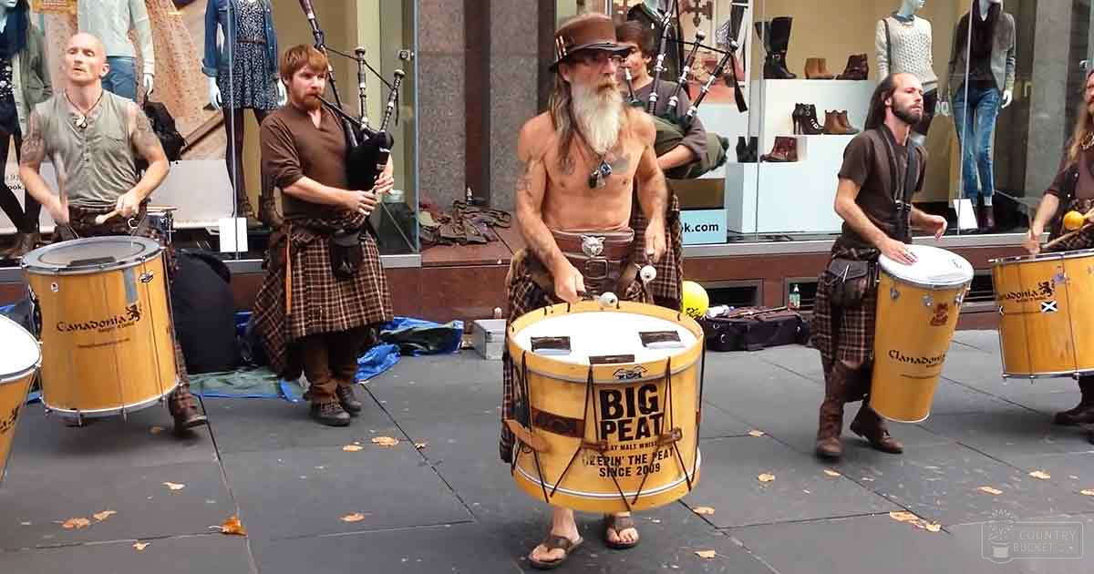 Street performance with an adult male singer in a patterned sleeveless shirt with dark pants; another man plays bagpipes wearing a brown tunic over a plaid kilt and boots, surrounded by musicians in similar attire with drums labeled 'Big Peat' and 'Clan Donald,' set against urban storefronts displaying mannequins dressed in clothing.