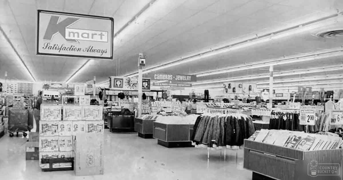Mid-20th century Kmart store interior with shelves stocked with cameras, jewelry, and denim jeans; fluorescent lighting fixtures overhead in a large retail space