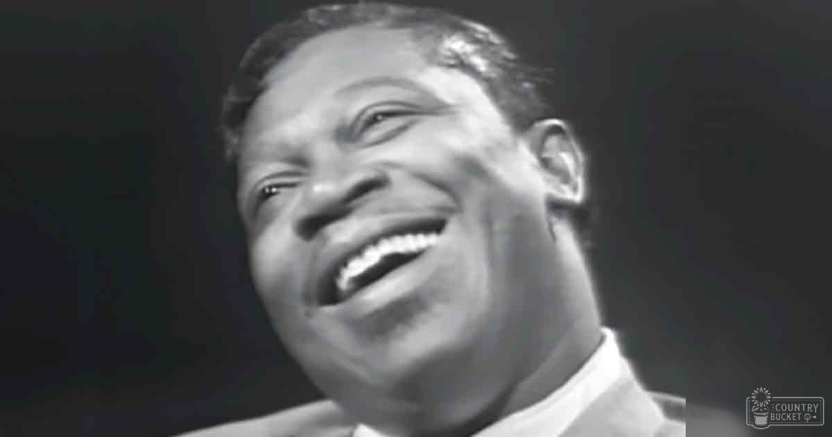 A smiling adult male wearing a light-colored collared shirt with classic fit, mid-20th century style, indoors under spotlight effect.