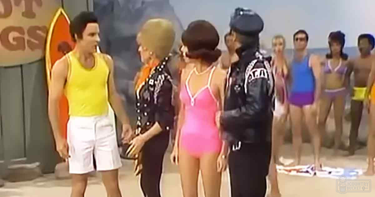 A group of people at a beach setting with sand and water visible in the background; adults wearing various styles of swimsuits, including a pink one-piece, black patterned suit, and yellow sleeveless top.