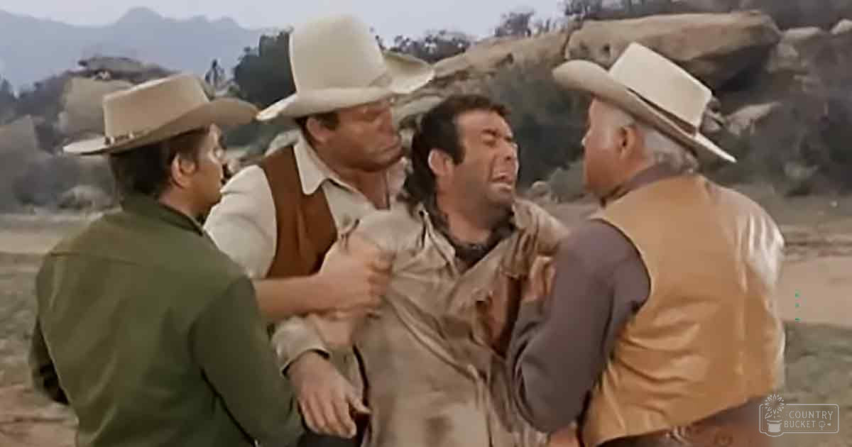 A group of men in a desert setting, likely from a Western film; adult males wearing wide-brimmed hats and jackets, appearing distressed or restrained.