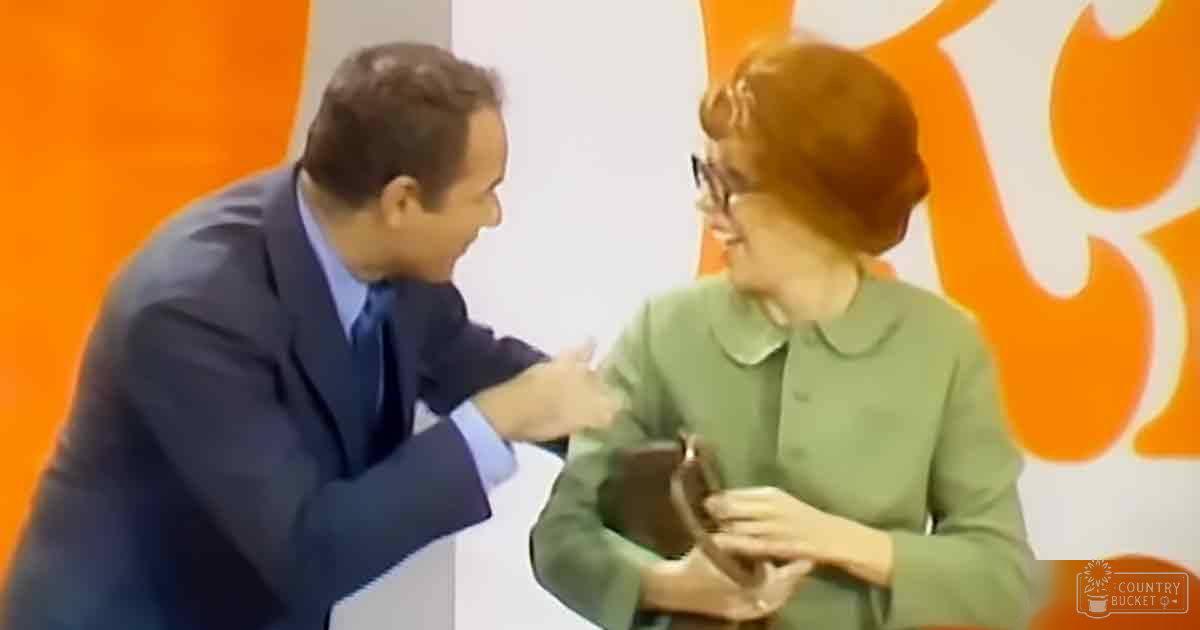 A man in a dark blue suit jacket and light blue dress shirt presents an object to a woman wearing a green collared top with long sleeves, set against an orange and white backdrop in what appears to be a professional studio environment.