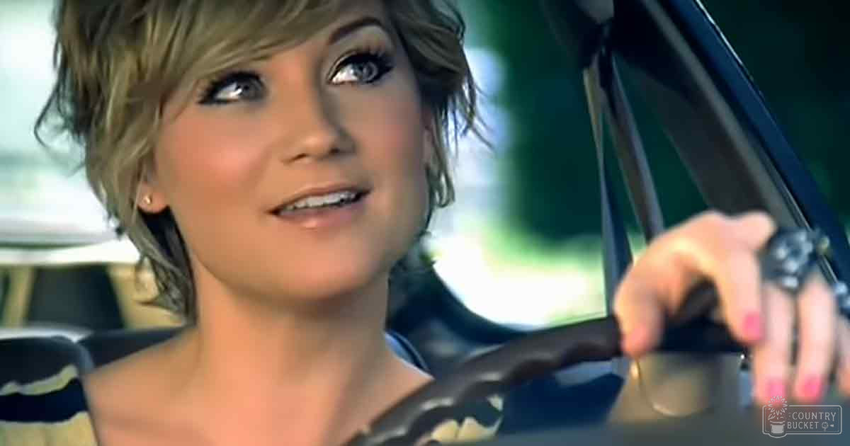 A young adult female drives a car in an outdoor setting with natural light filtering through trees.
