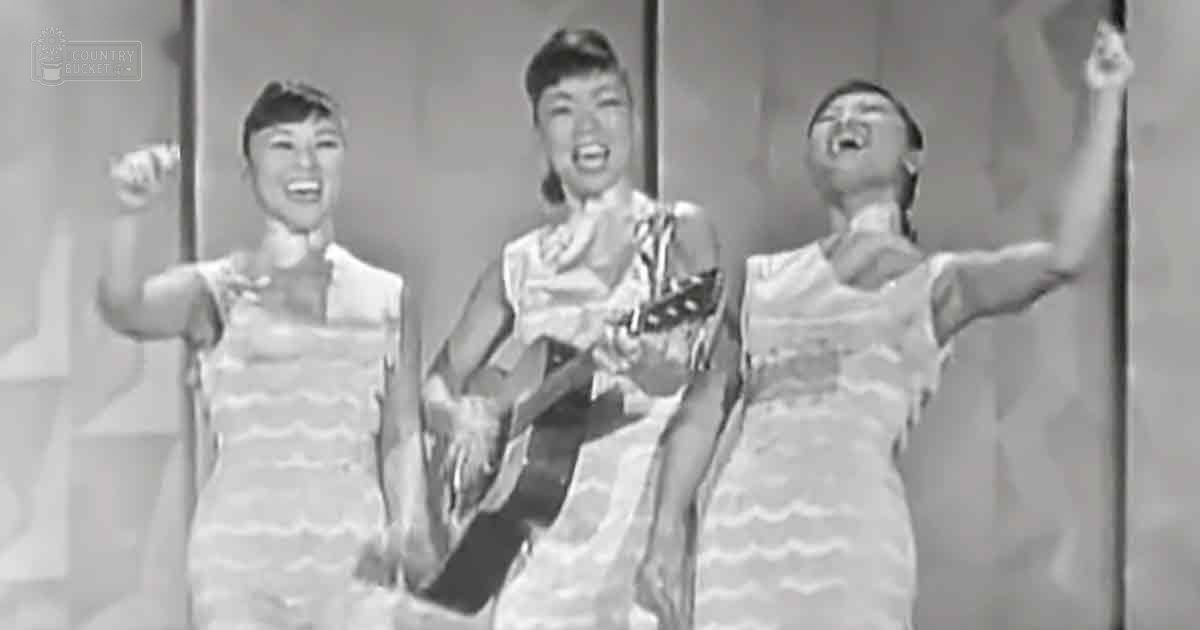 A musical performance scene with young adult females in sleeveless, striped dresses against a simple studio-like background.