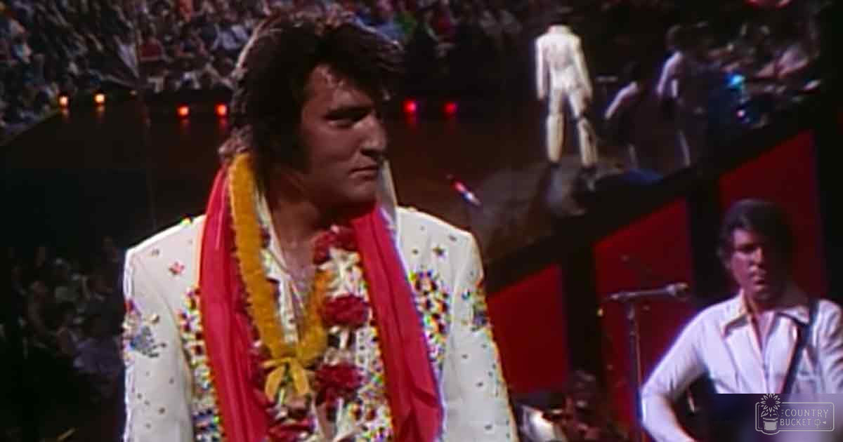 An adult male performer wearing a white sequined jacket with floral patterns and red and yellow leis, accompanied by another young adult male in a light-colored shirt on stage during a concert.