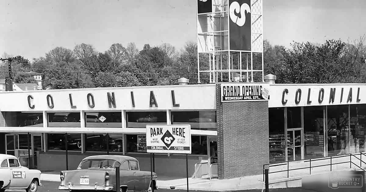 Vintage cars parked on urban street with 'Park Here' sign, large windows of store advertising grand opening soon, bare trees suggesting late fall or early spring, daytime setting in black and white image.