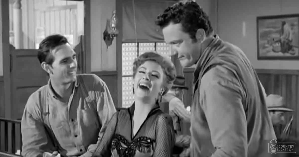 An adult female seated at a bar or saloon setting with rustic Western decor, wearing a patterned dress over a lighter top; an adult male on her left wears suspenders and a light shirt, suggesting workman's attire.