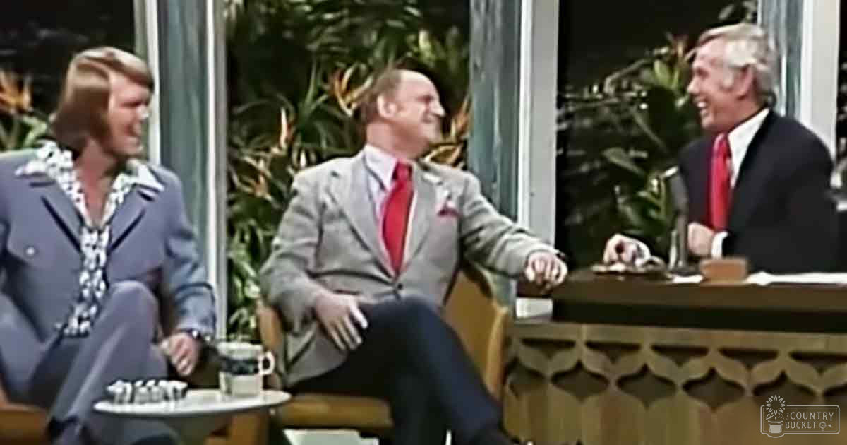 Three individuals seated around a small round table in a talk show setting; adult female wears patterned blouse and gray blazer, middle-aged male in light gray suit with red tie, older adult male in dark suit with red tie. Indoor studio with large windows showing greenery outside.