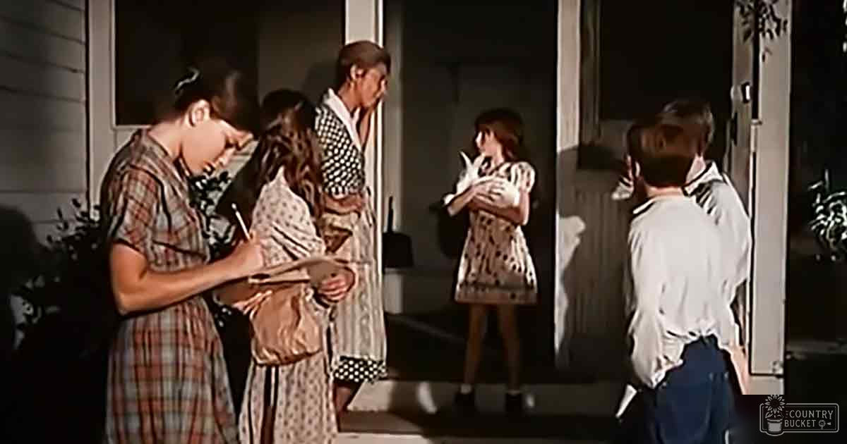Children in plaid dresses interacting with adults on a residential porch at night, surrounded by artificial lighting and hints of summer greenery.