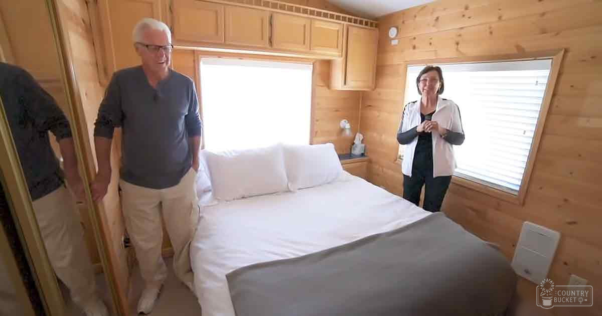 A middle-aged man and an older adult woman are positioned indoors in a bedroom with rustic wooden paneling; they wear casual attire with muted colors, natural light fills the space from windows with blinds partially open.