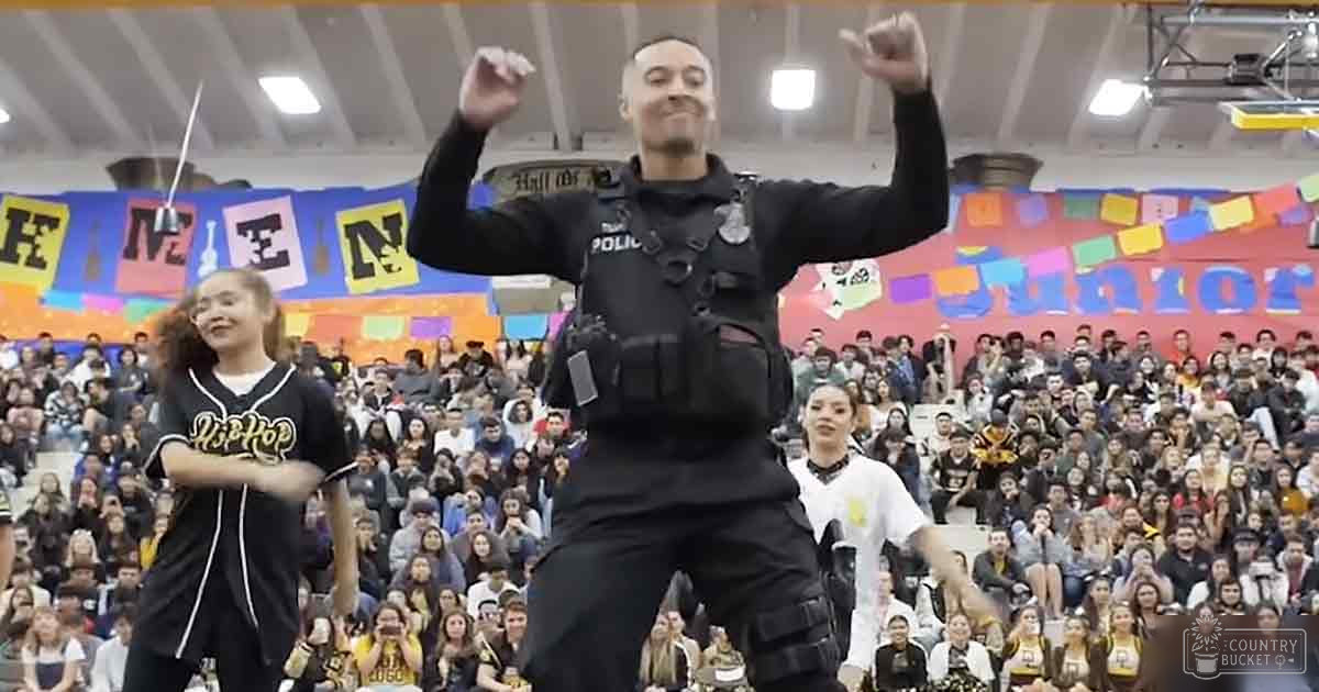 A police officer wearing a black tactical uniform with 'POLICE' written on it performs in front of an audience at a school gymnasium, alongside other individuals dressed casually and actively participating.