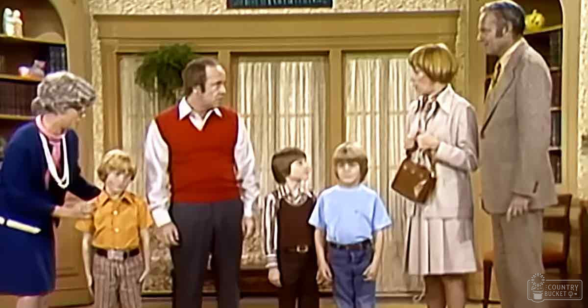 Family scene indoors with adults and children wearing various outfits including a dark blue dress with white trim and pink blouse, red vest over white shirt, striped brown and white long-sleeve shirt. Bookshelves and potted plant in the background suggest a cozy living space.