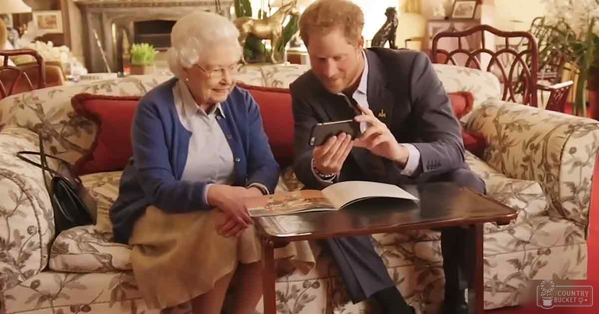 An older adult woman wearing a light-colored blouse with long sleeves, a blue cardigan, and a beige skirt or dress, alongside a young adult man in a dark suit jacket holding a smartphone, in a cozy living room setting with floral-patterned furniture and decorative elements.