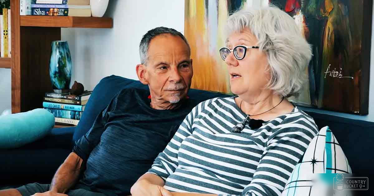 Older adult couple in a cozy living room with blue sofa, striped shirts, warm-toned painting, and natural light.