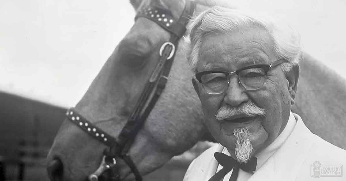 Colonel Sanders in a white suit jacket with a black bow tie, standing next to a light-colored horse on an open farm setting under natural daylight.