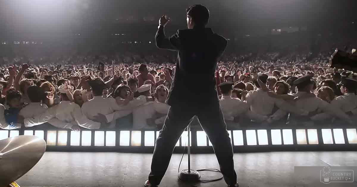 Children in white uniforms with caps seated on bleachers at a lively concert or event indoors under bright artificial lighting.
