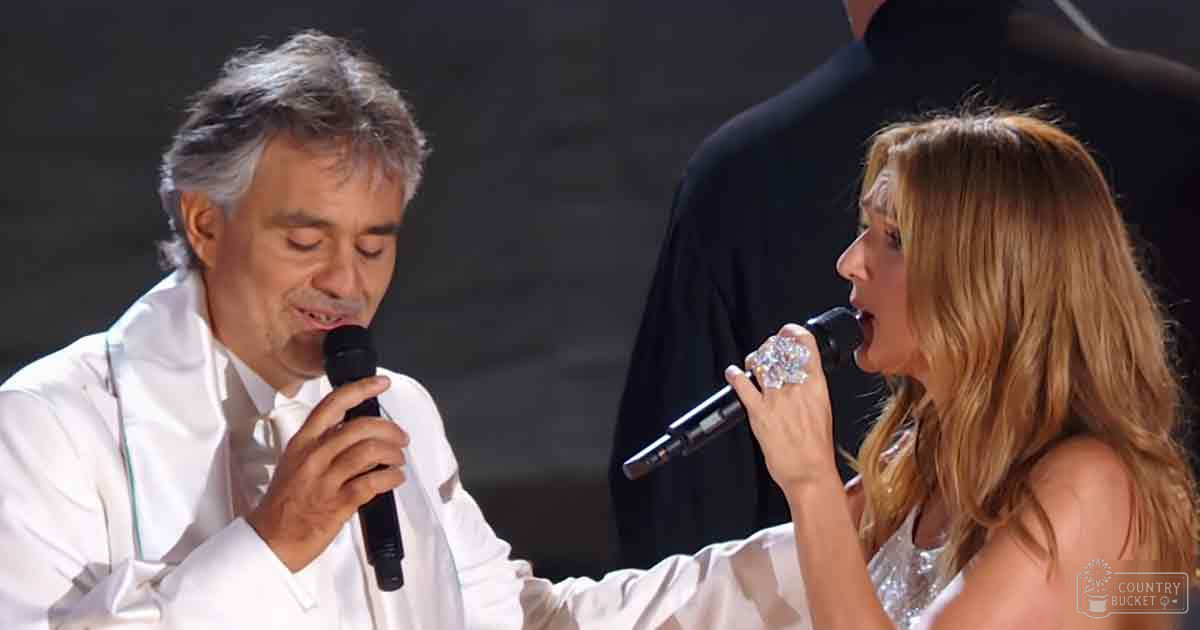 A duet performance scene at an indoor concert venue with stage lighting, with two individuals holding microphones; one middle-aged man wearing a white satin garment and another young adult woman in a shiny top or dress.