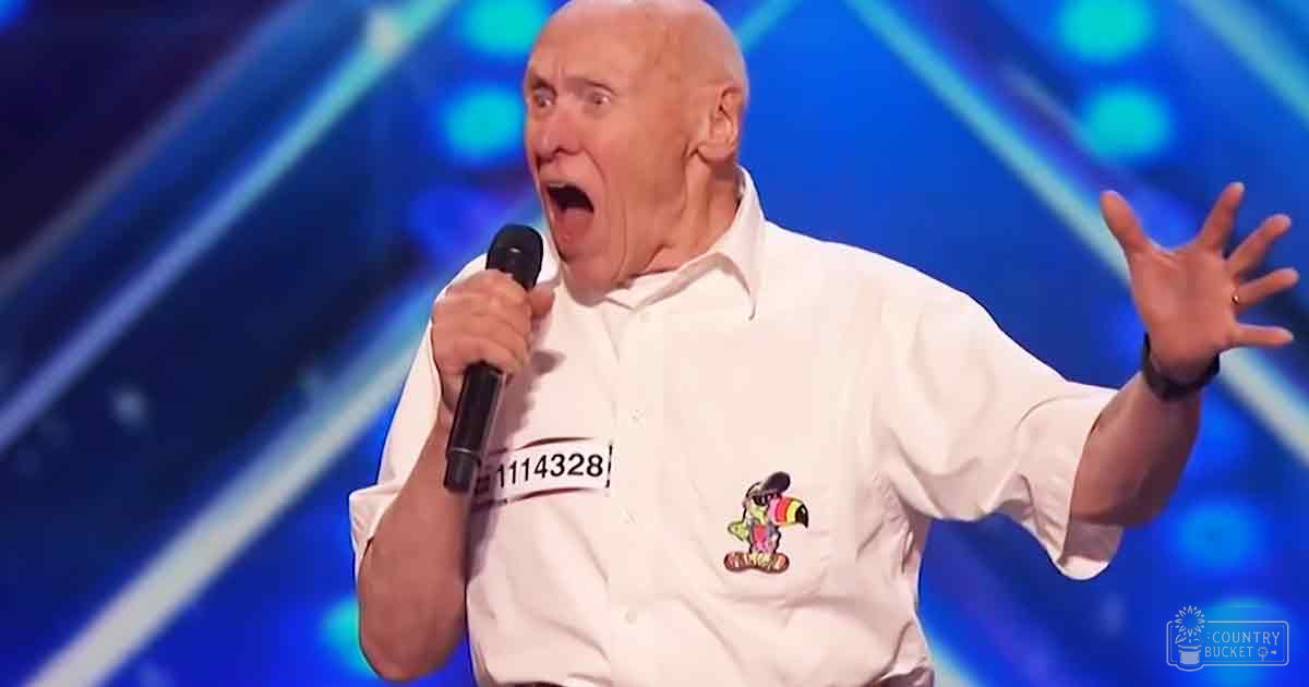 An older adult male in a white polo shirt with a graphic design and number '1114328', performing on stage, holding a microphone.