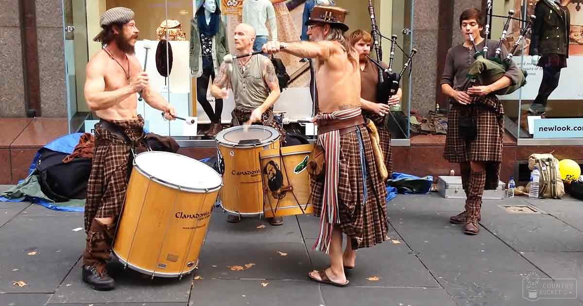 A street performance group in an urban setting, with adults wearing plaid outfits and historical attire, with drums labeled 'Clanadonia' prominently displayed, set against a backdrop of mannequins dressed for cooler weather under natural daylight.