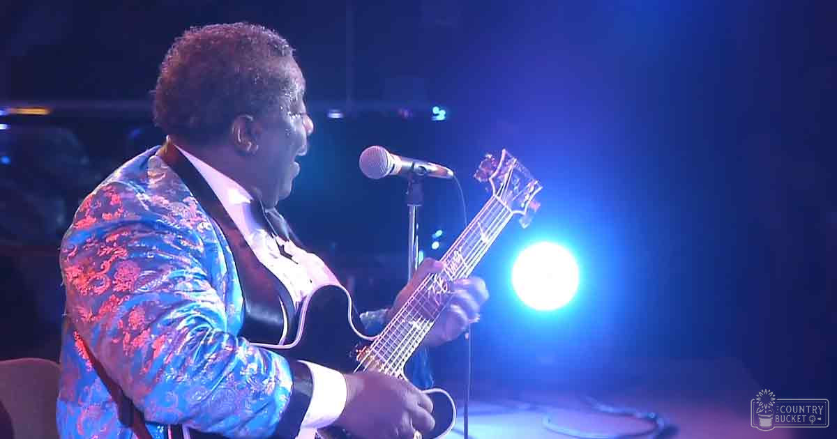 A middle-aged to older adult male singer in a vibrant, patterned floral jacket plays guitar on stage at an indoor concert venue with blue stage lighting and 'Madly Odd' text printed nearby.