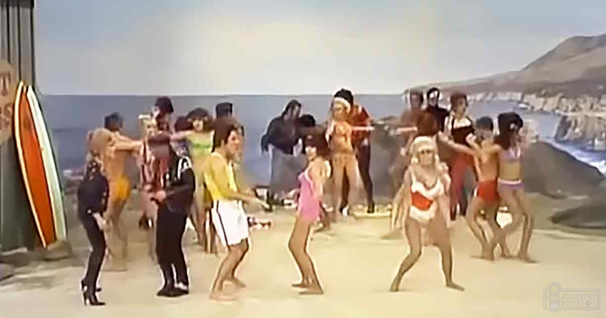 Beach scene with group of people dancing; staged or filmed beach setting; adults and children in summer attire; painted backdrop depicting sand, water, distant hills under clear sky; daytime natural sunlight; various light blue, white, blue, beige, and colorful clothing tones